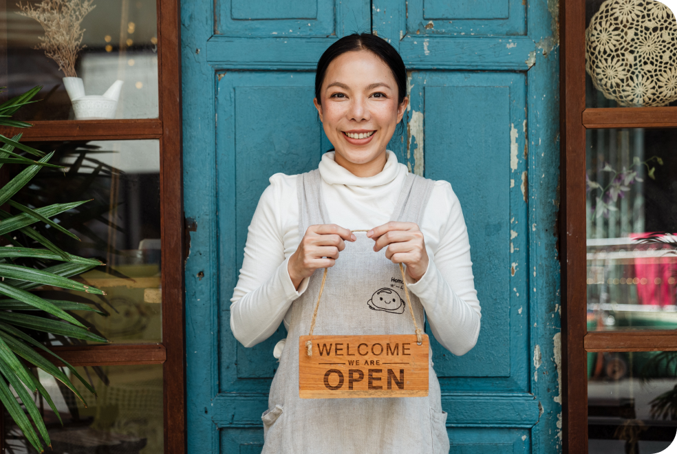 A woman is smiling and holding a wooden open sign in front of the restaurant.