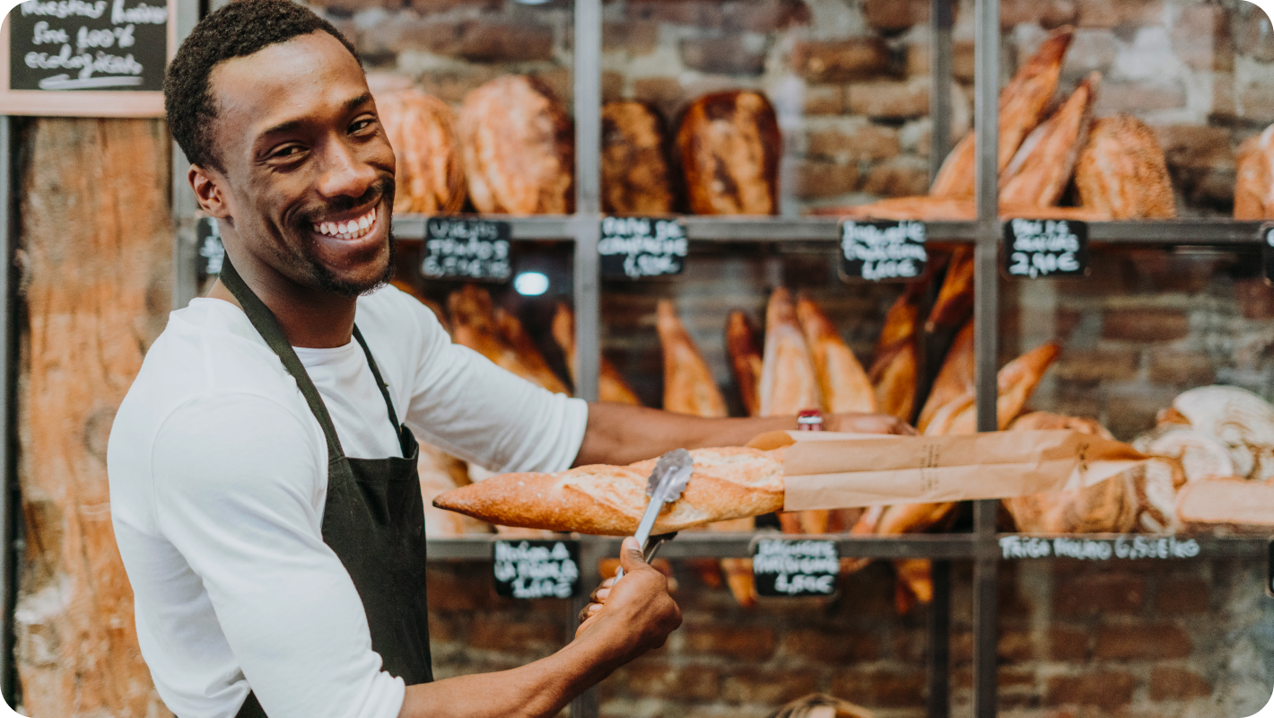 An employee standing in the bakery shop is holding warm bread.