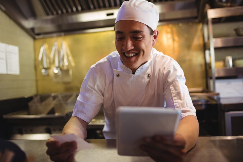 A chef is holding an order receipt and a tablet in the kitchen.