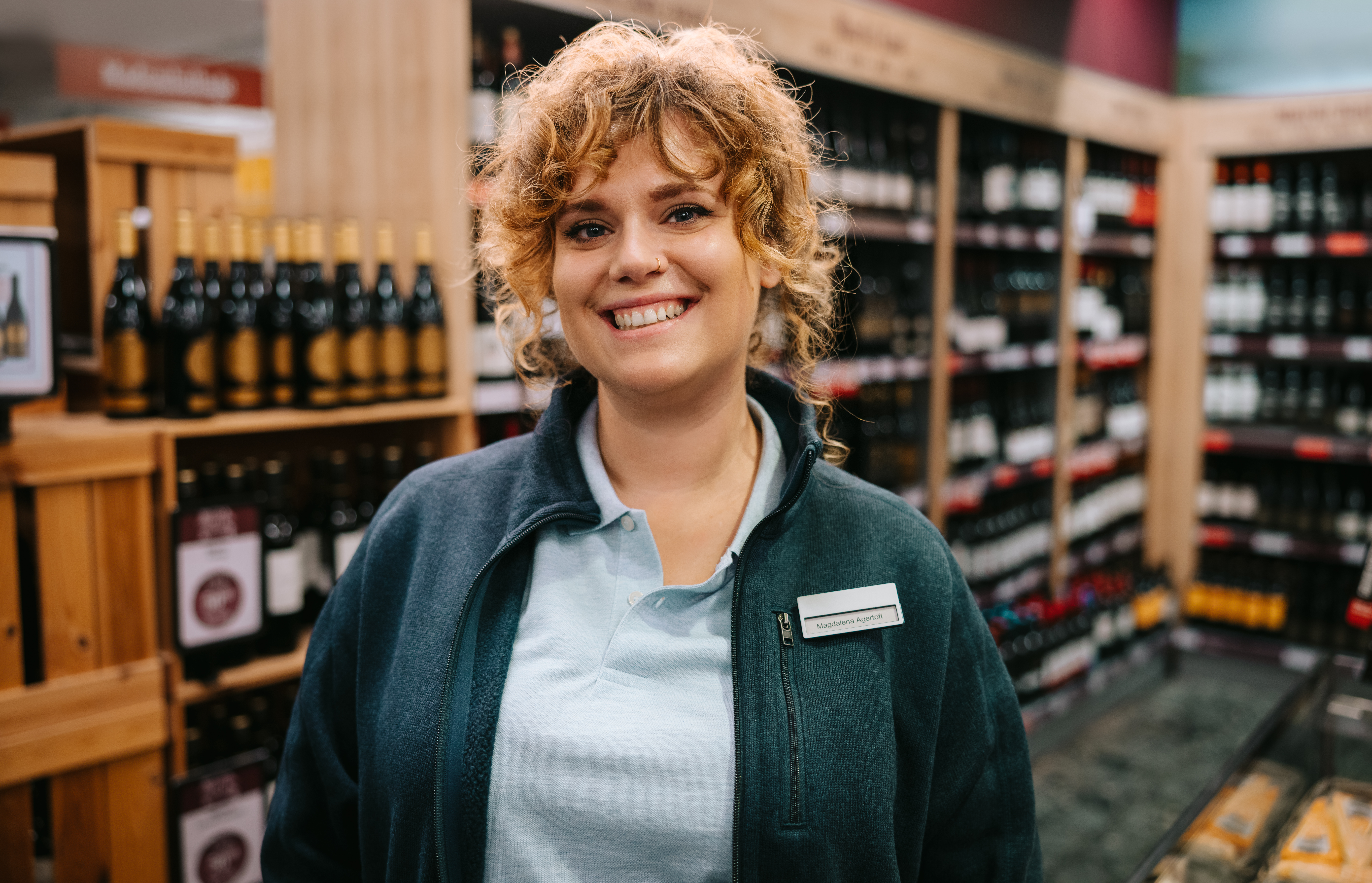 A female employee standing in the liquor store is smiling.