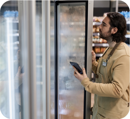 An employee standing in the convenience store is opening the fridge to check the inventory while holding the tablet.