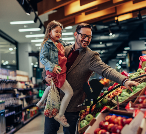 A man holding his daughter in his one arm is picking fruit from the grocery store fruit section.