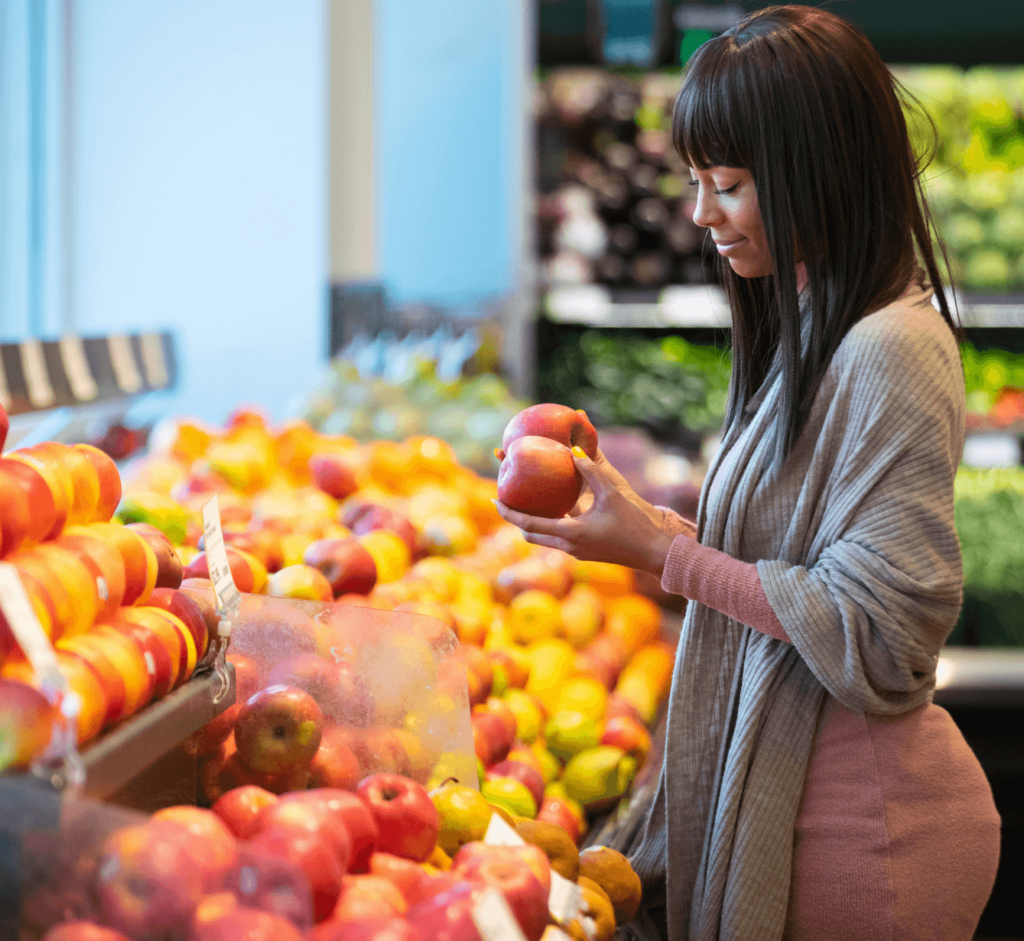 A woman standing in the fruit section is holding two apples in her hand.