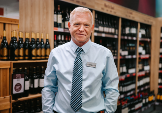 A liquor store manager wearing a business shirt and tie is standing in the liquor section.