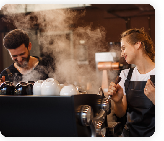 A male and female employee standing in the cafe kitchen are using a coffee roaster machine.