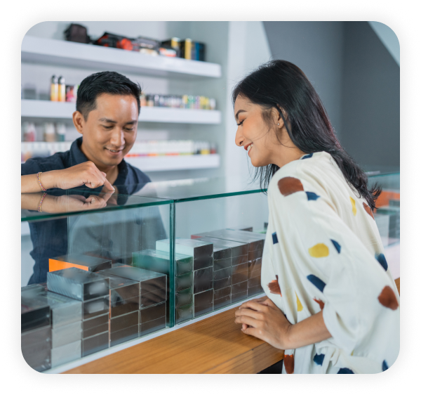 A smoke shop employee standing at the checkout counter is assisting a female customer in choosing the vape.