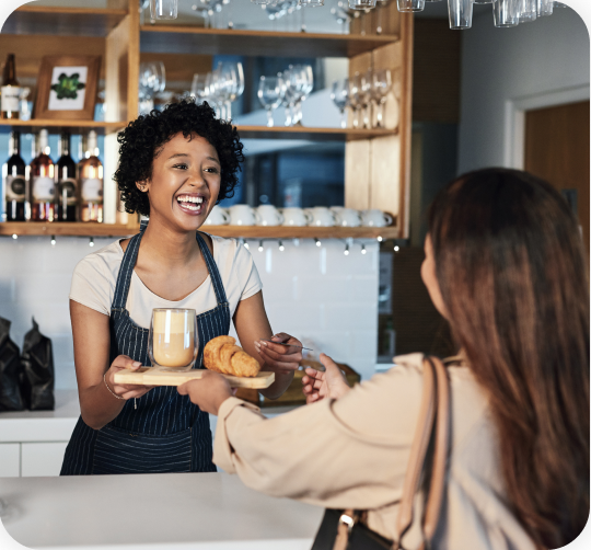 A female employee wearing an apron is providing coffee and a loaf of bread to the female customer.