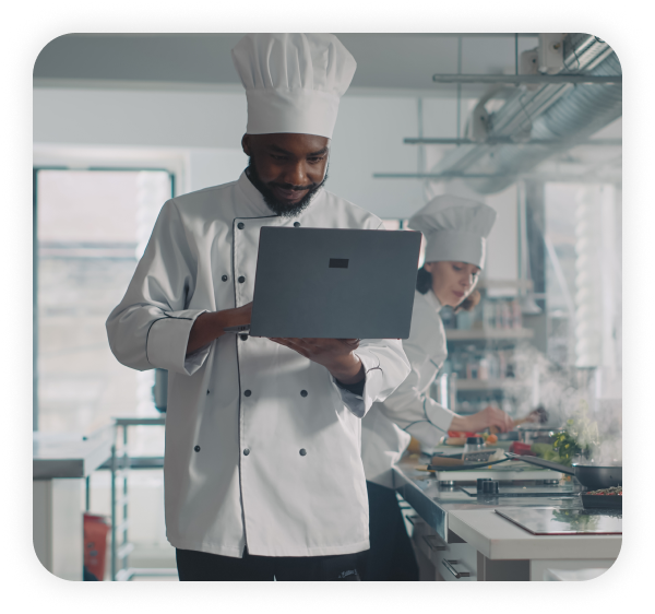 A chef standing in the kitchen is using inventory management system software on the laptop.