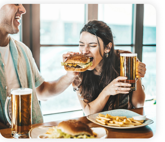 A young girl sitting in a restaurant is taking a burger bite while holding a beer.