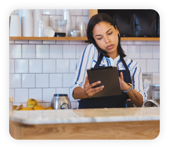 A female employee wearing an apron standing in the fast-food restaurant kitchen is using a tablet while attending a call.