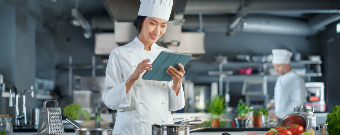 A female chef standing in the kitchen is using a tablet.