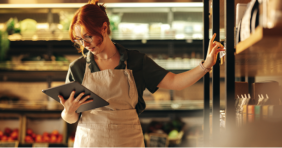Store employee reviewing inventory on a tablet beside refrigerated shelving in a retail food environment.