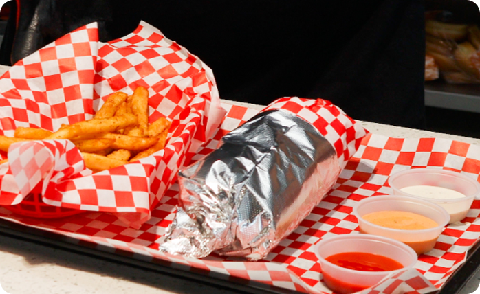 Food service meal featuring a wrapped burrito, seasoned fries, and dipping sauces served on a tray.