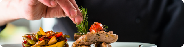 Close-up of a chef adding fresh rosemary garnish to a plated entrée with vegetables, mushrooms, and roasted ingredients.