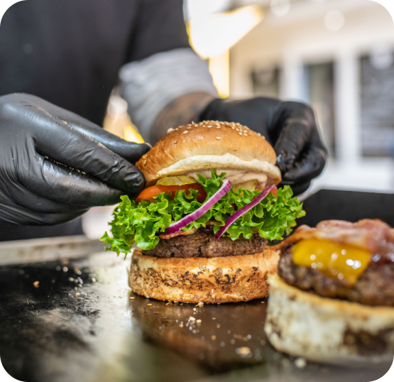 Food service worker wearing black gloves assembling a gourmet burger topped with lettuce, red onion, and sauce on a toasted bun.
