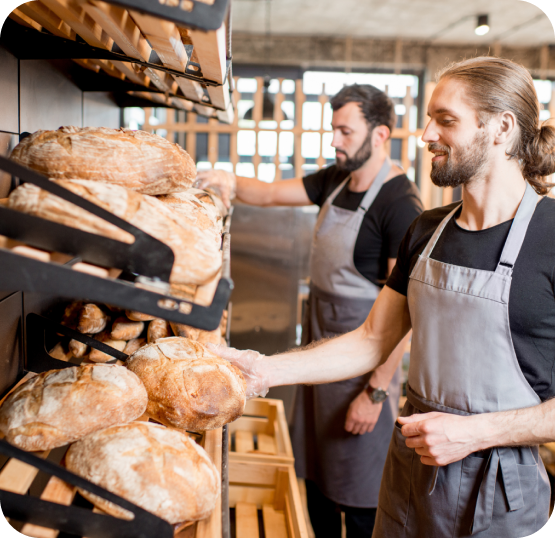 Bakery staff stocking shelves with freshly baked artisan bread loaves, displayed on wooden racks inside a commercial bakery setting.