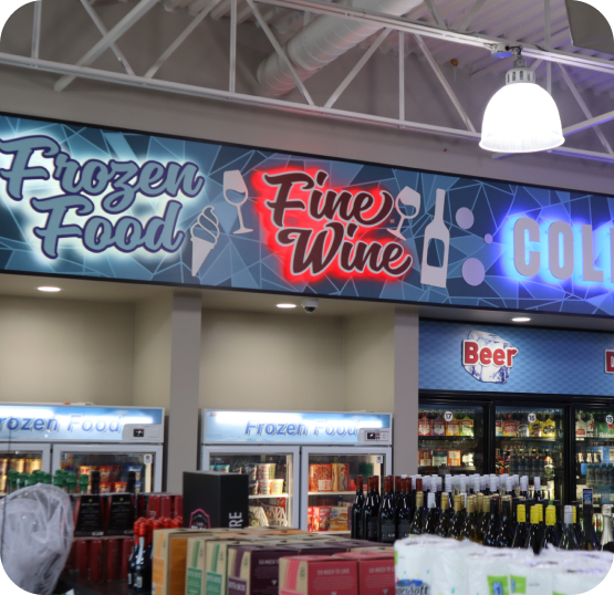 Convenience store beverage section featuring illuminated Frozen Food, Fine Wine, and Beer signage above refrigerated cases and stocked shelves.