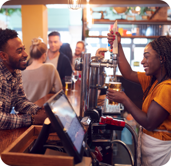 Bar staff serving draft beverages to customers at a bar counter with a point of sale workstation nearby.