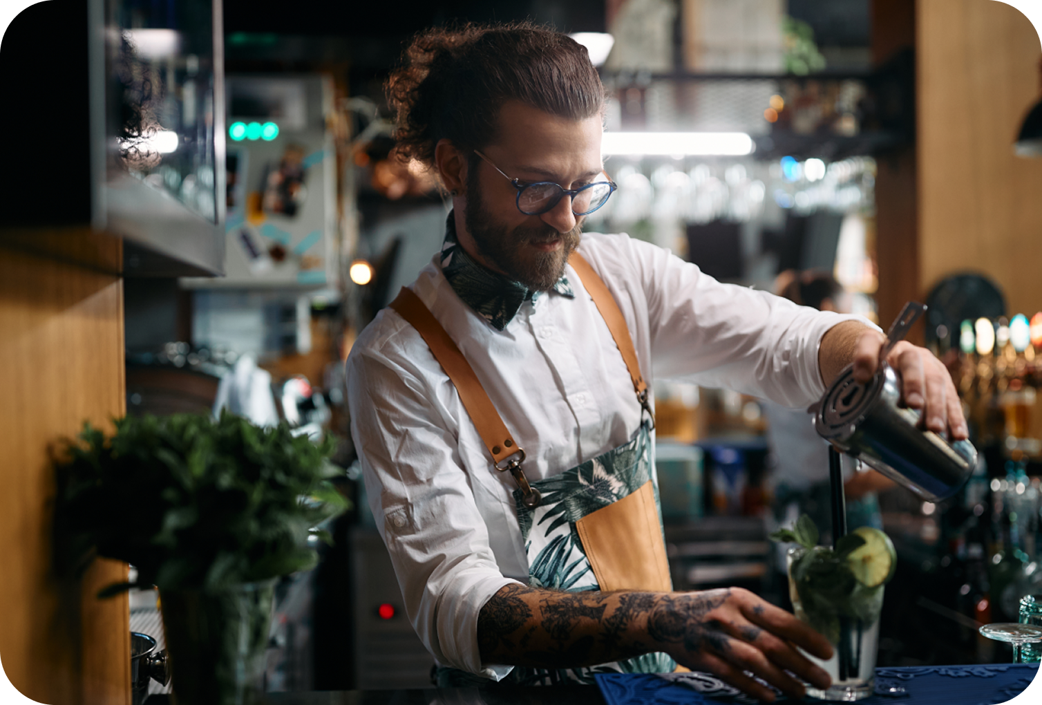 Bar staff member preparing a cocktail behind a bar counter while pouring a beverage into a glass garnished with lime slices.