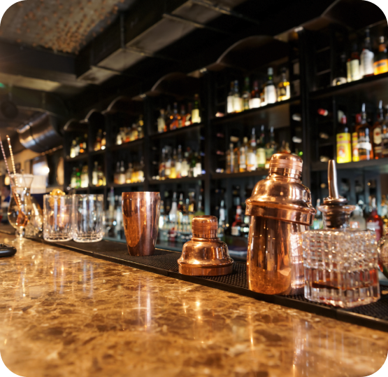 Bar counter displaying cocktail shakers, glassware, beverage tools, and liquor bottles arranged on shelving in the background.