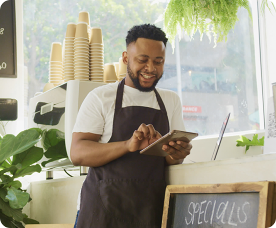 Cafe employee using a tablet behind a service counter with takeaway coffee cups, menu displays, and bakery service equipment in the background.