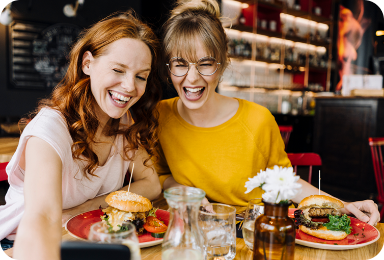 Guests seated at a restaurant table with burgers, beverages, and tableware while viewing content on a mobile device.
