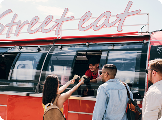 Customers collecting food orders from a food truck service window while a staff member hands over a prepared meal.
