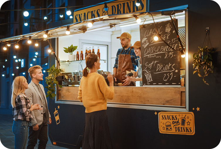 Customers placing and receiving food orders at a food truck service counter illuminated by outdoor string lights.