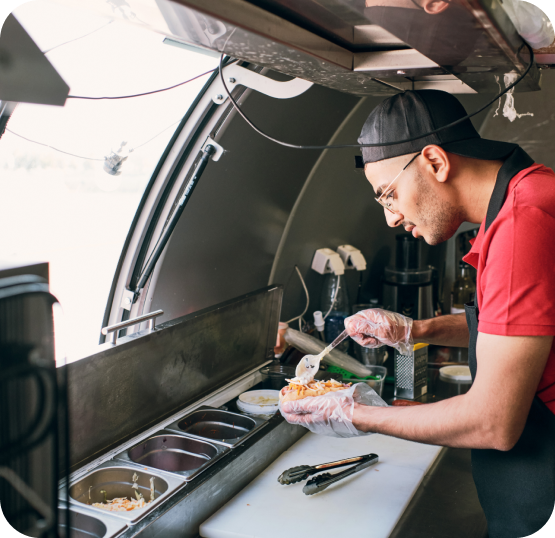Food service worker preparing meal ingredients at a food truck workstation with food containers, utensils, and preparation equipment.