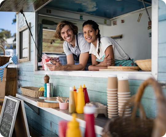Food truck staff leaning from a service window above a counter with condiment bottles, disposable cups, and food service supplies.