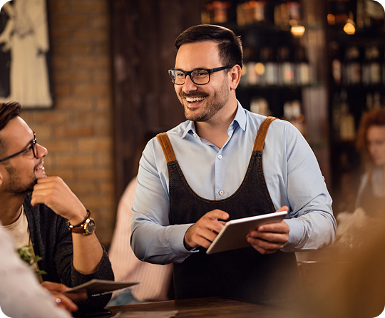 Restaurant staff member using a tablet to take an order while interacting with guests at a dining table.