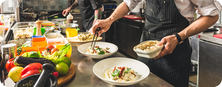 Kitchen staff plating prepared dishes at a commercial restaurant workstation with fresh ingredients, cooking equipment, and food preparation stations.