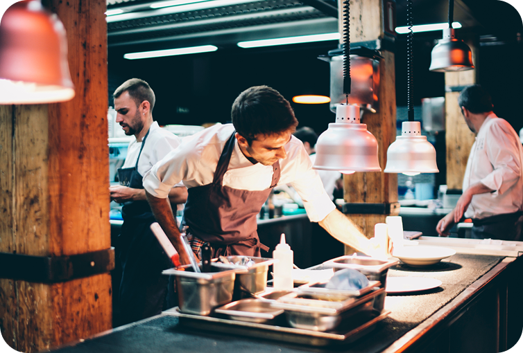 Kitchen staff preparing food orders at a commercial restaurant cooking station with heat lamps and food preparation equipment.